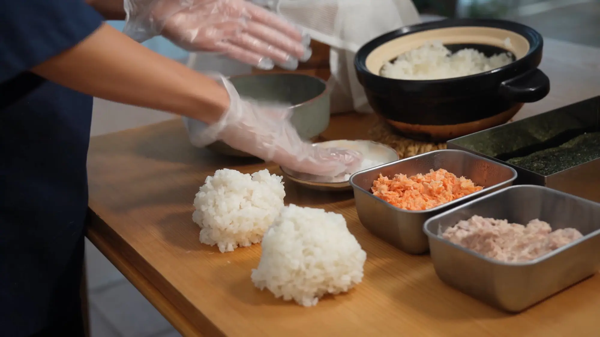 Gloved hands taking a pinch of salt from a small dish onto damp palms before shaping rice