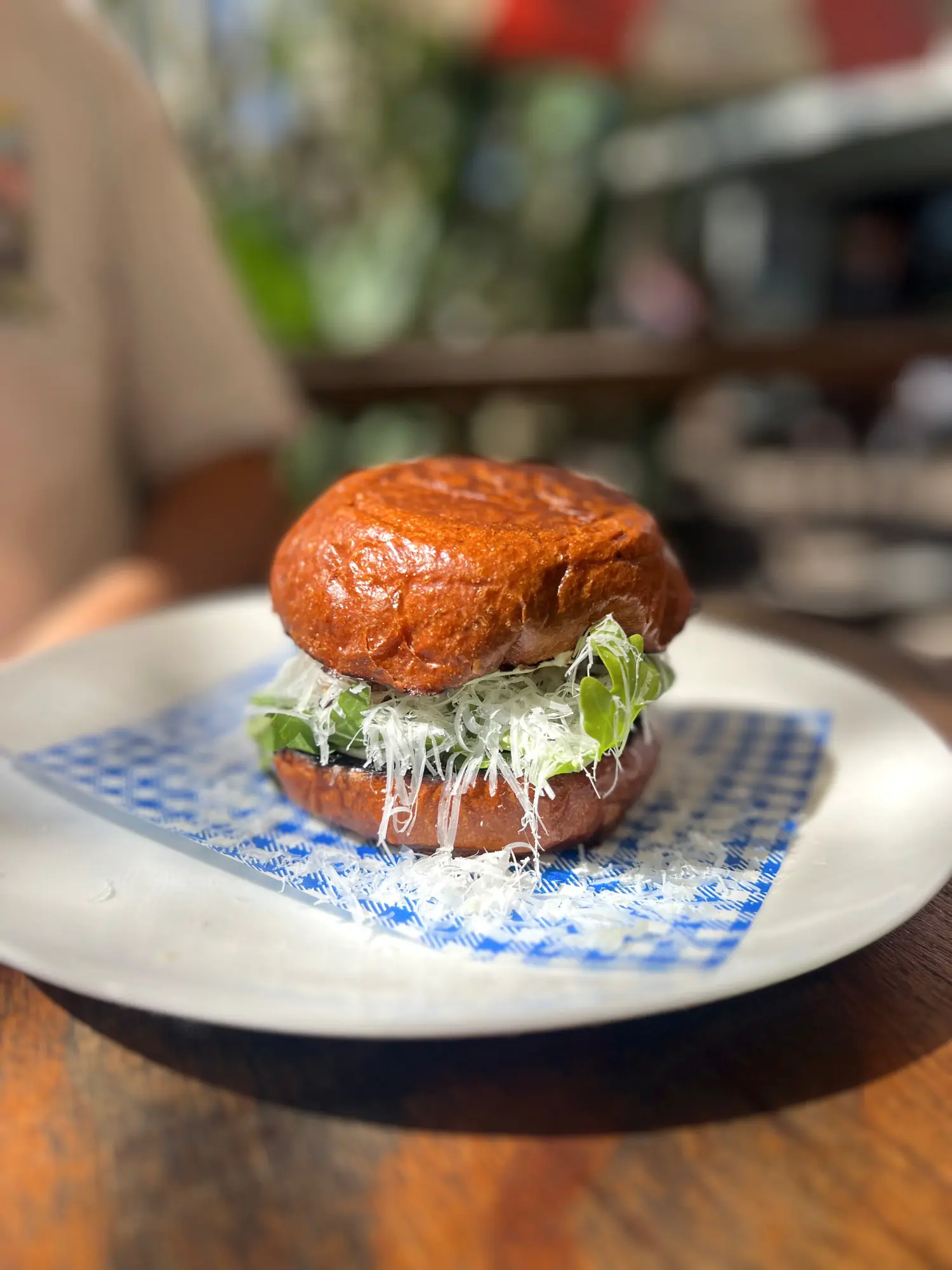 A glossy brioche burger topped with greens and finely grated cheese on a blue-checked wrapper at The Salty Mangrove