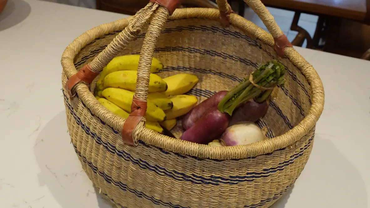 A woven market basket at home filled with bananas, purple sweet potato, turnips and asparagus