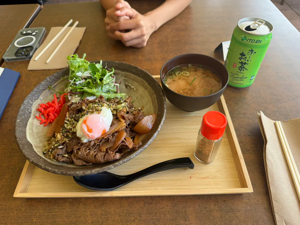A gyudon beef rice bowl with a soft onsen egg, furikake, red pickled ginger and salad on a stone bowl with miso soup and a green tea can at Ebiya
