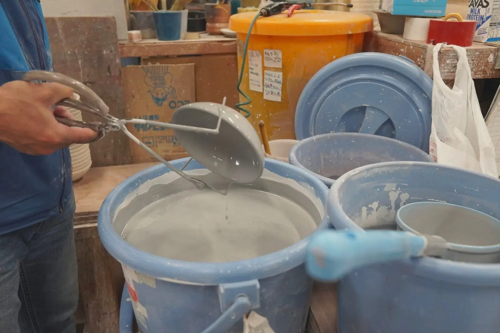 A craftsman holding a lid with tongs and dipping it into a bucket of grey glaze.