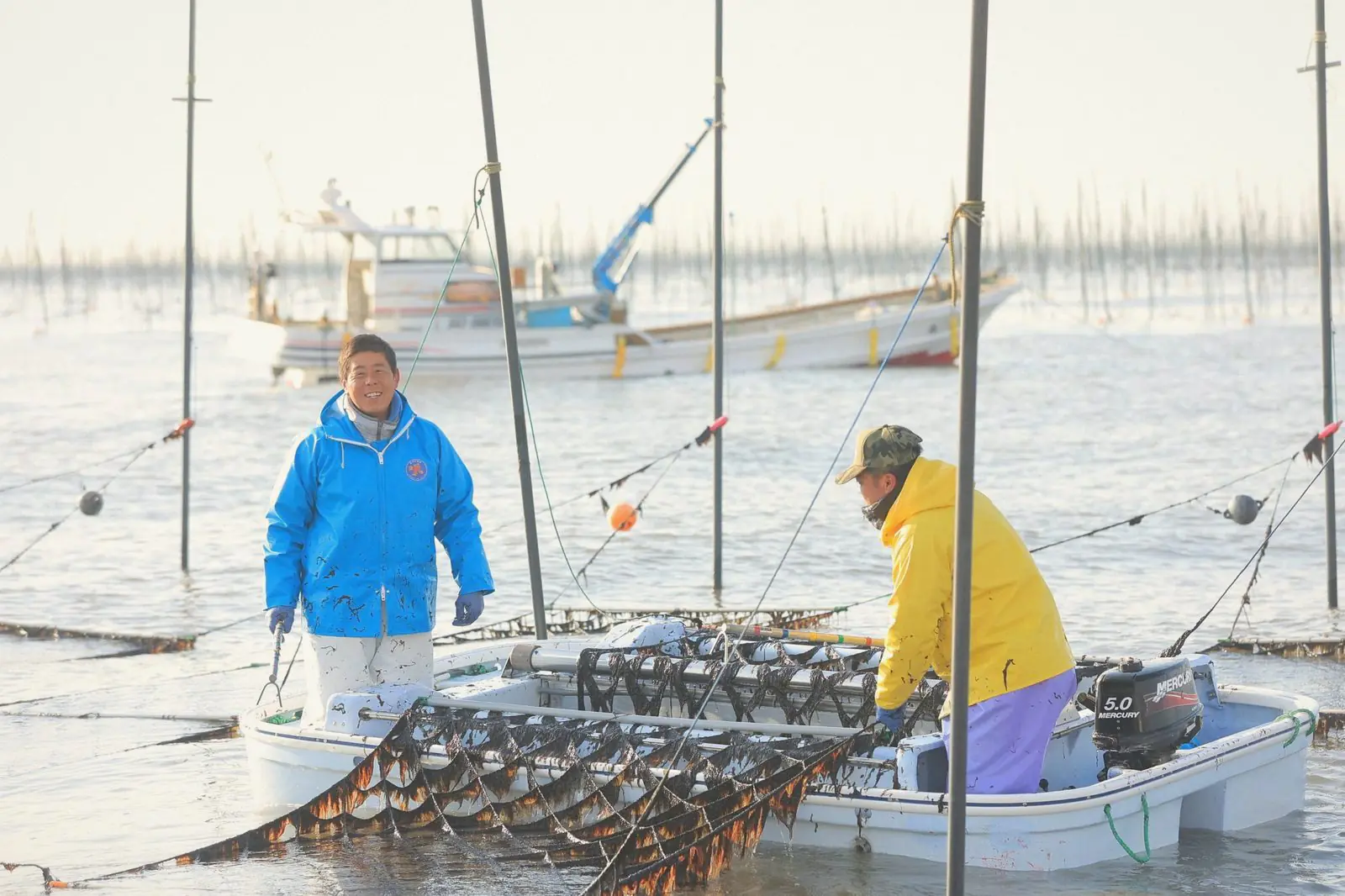 A smiling nori farmer in a blue jacket standing in the water beside a harvesting boat.