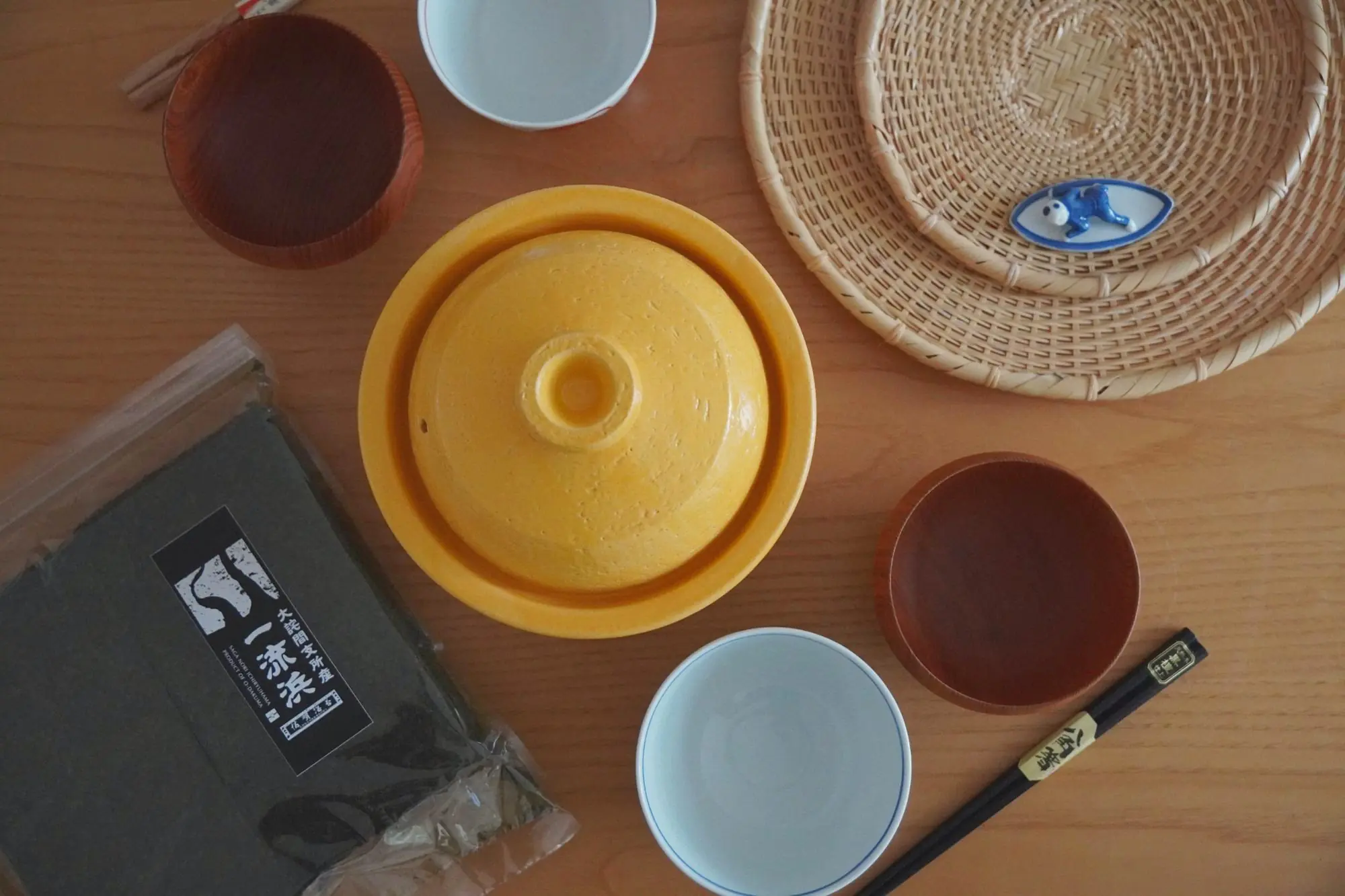 A mustard Matsusho donabe on a wooden table with Ichiryūhama nori, rice bowls, lacquered bowls and chopsticks.