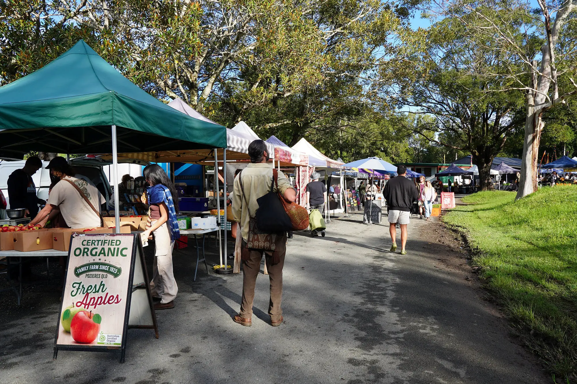 Shoppers walking the tree-shaded aisle of the Mullum Farmers Market past a Certified Organic fresh apples sign