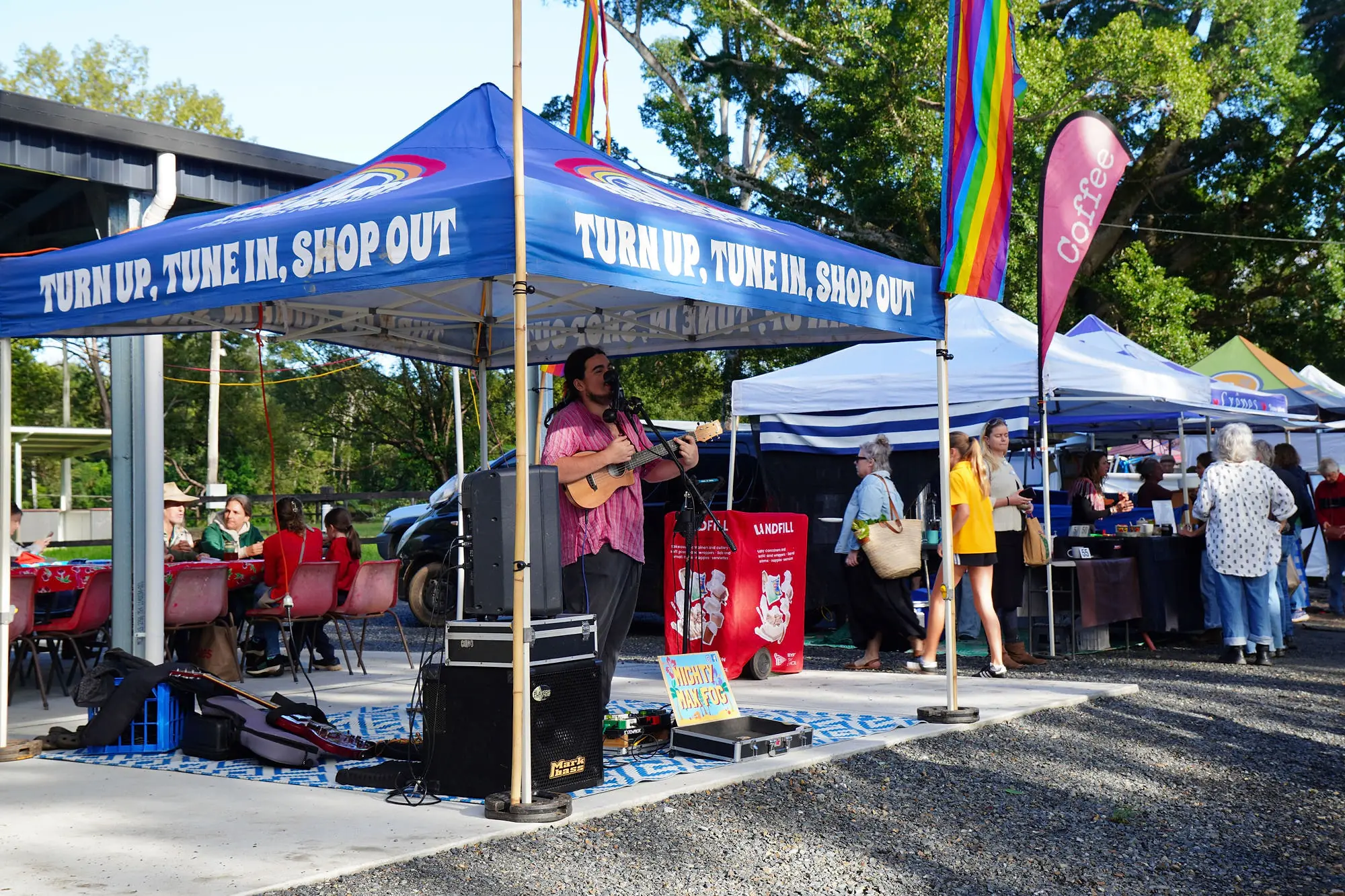 A musician playing live under a market gazebo reading 'Turn up, tune in, shop out' at the Mullumbimby Farmers Market