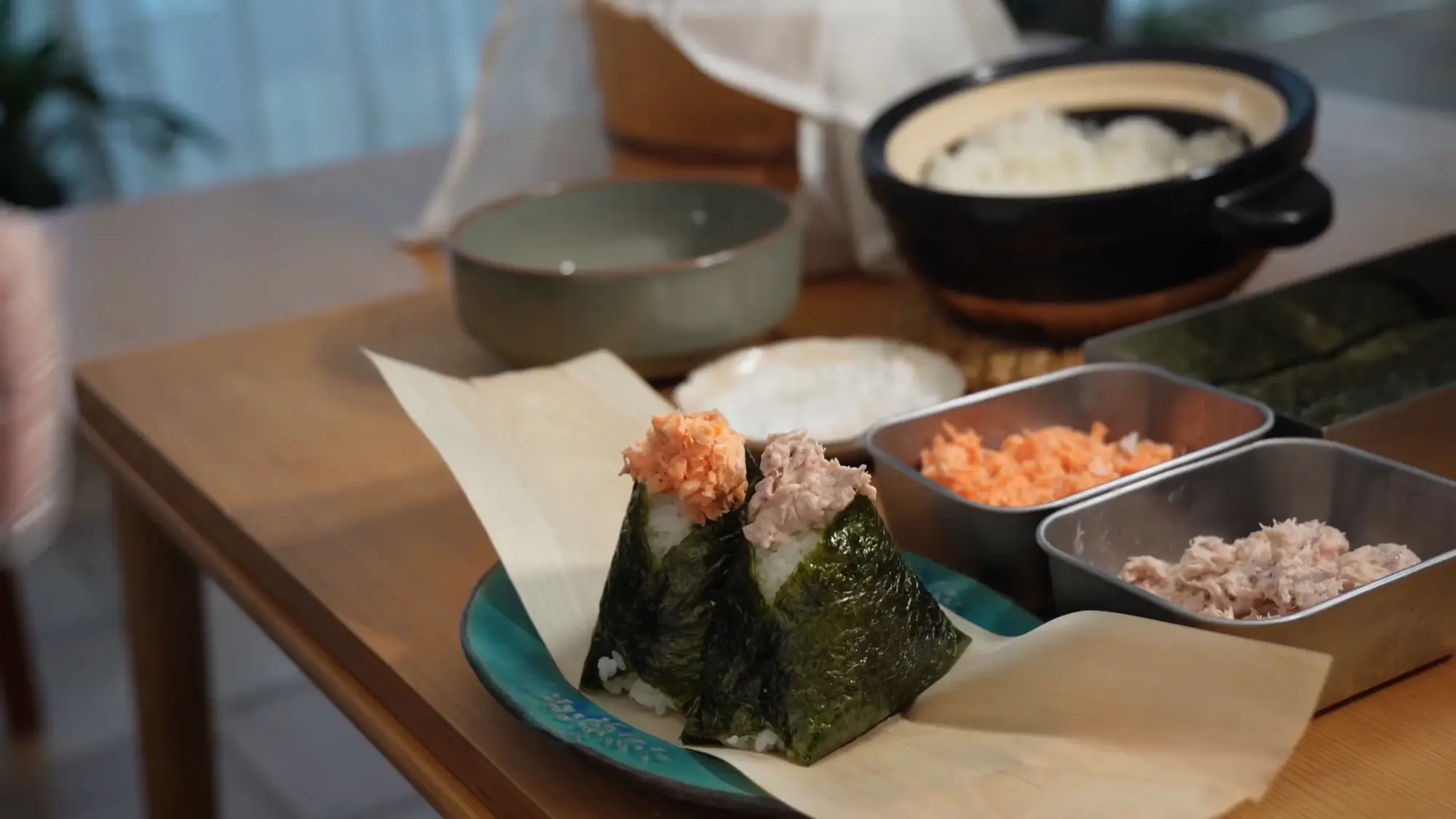Two finished onigiri — one topped with flaked salmon, one with tuna — wrapped in dark nori, beside a clay pot of rice and trays of fillings