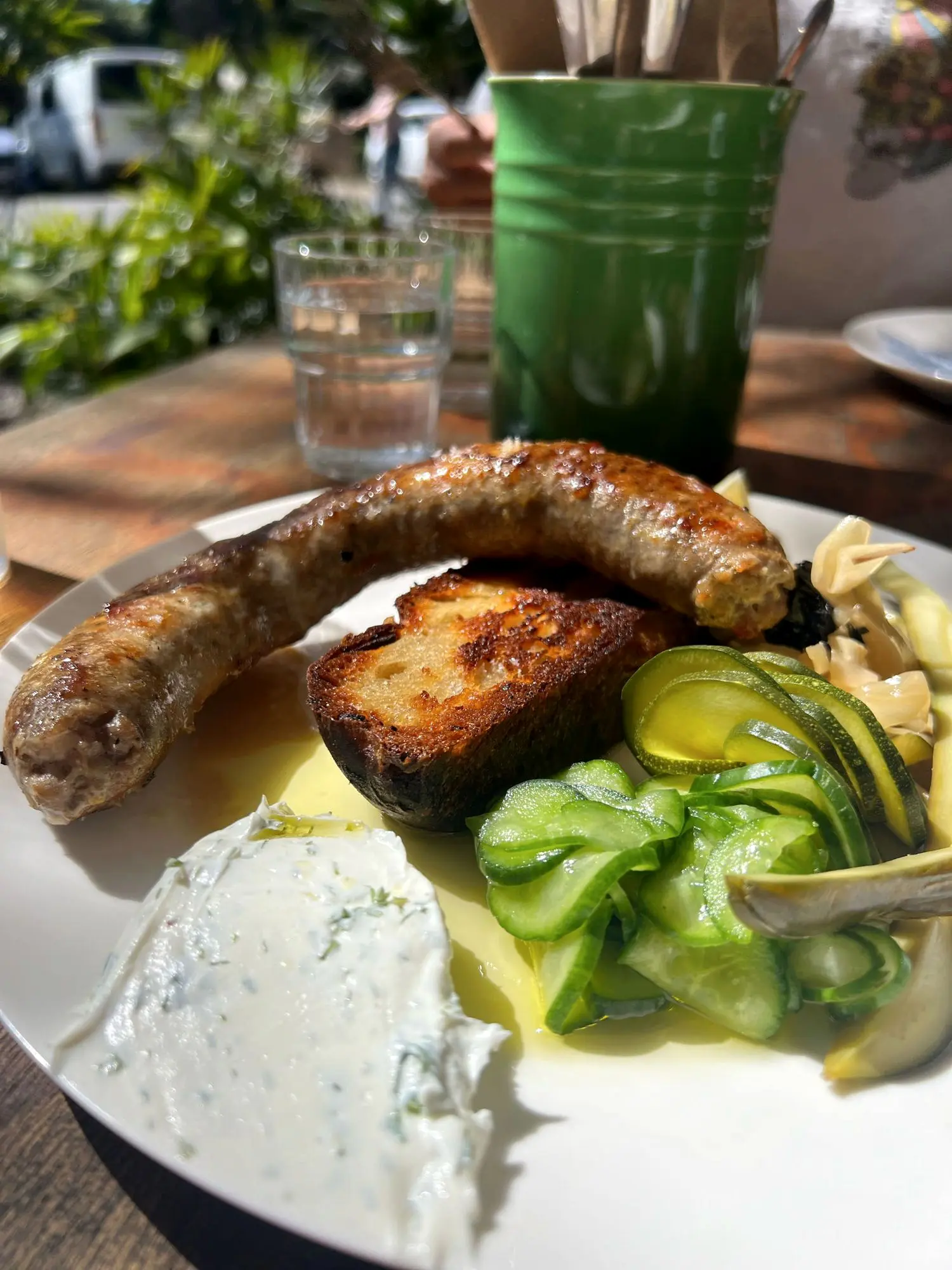 A grilled sausage with charred sourdough, pickled cucumber and a herbed dip on a plate in the sun at The Salty Mangrove